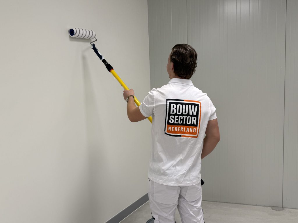 Schilder in wit werkpolo rolt met een telescoopsteel een lichte wand in een lege ruimte