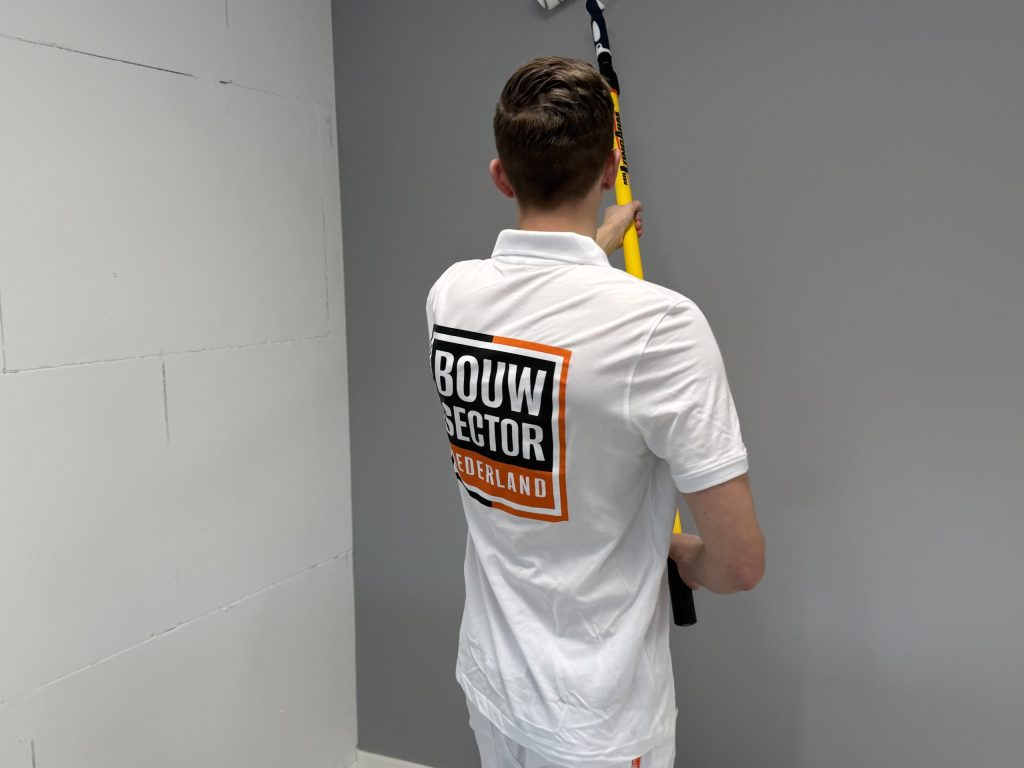 Schilder zet de roller hoog tegen een grijze muur naast een kale lichte wand