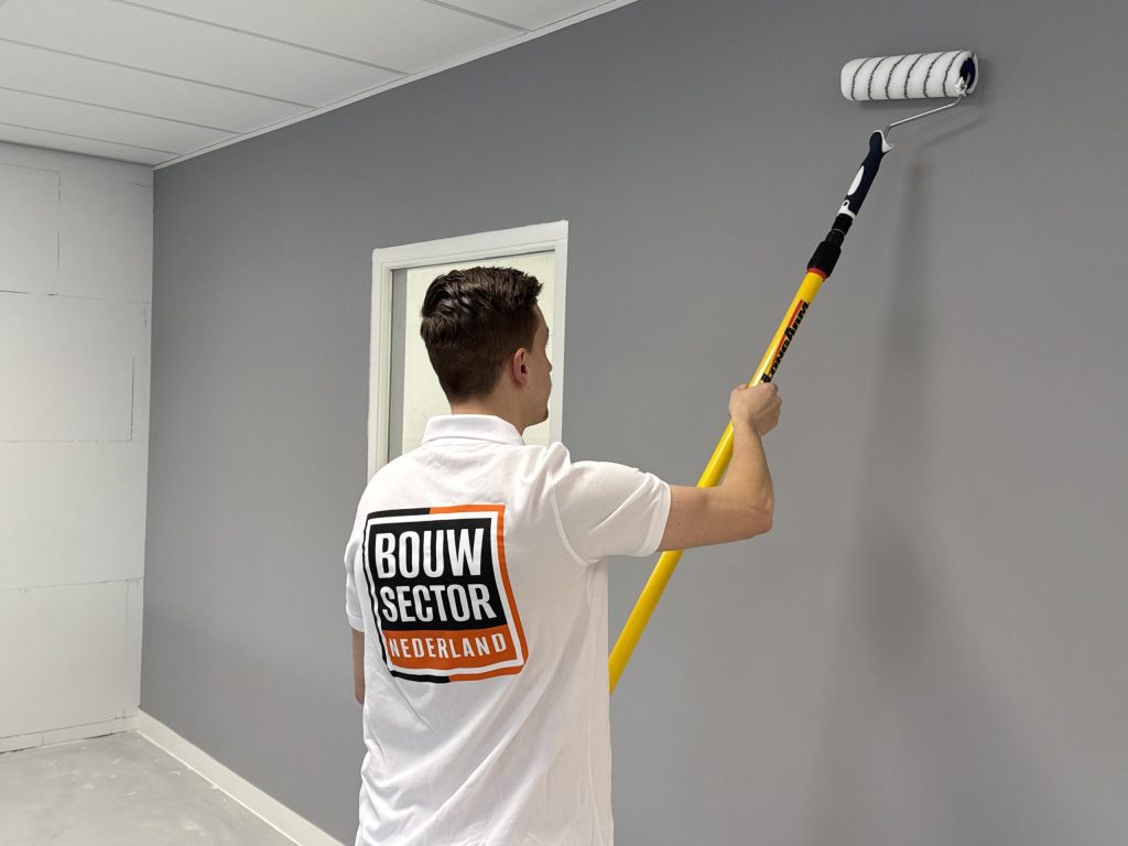 Schilder in wit polo rolt grijze verf op een muur naast een witte deuropening